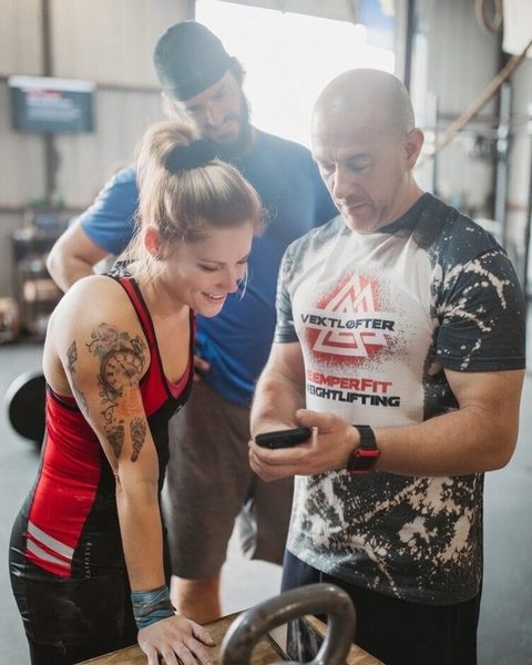BoxBestie founder Jim Roncal shows an athlete the BoxBestie app on his phone on the gym floor as another athlete looks on.